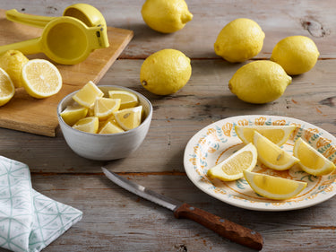 A selection of Eureka lemons on a wooden surface, with one being cut into slices on a plate, alongside a lemon squeezer and a knife.